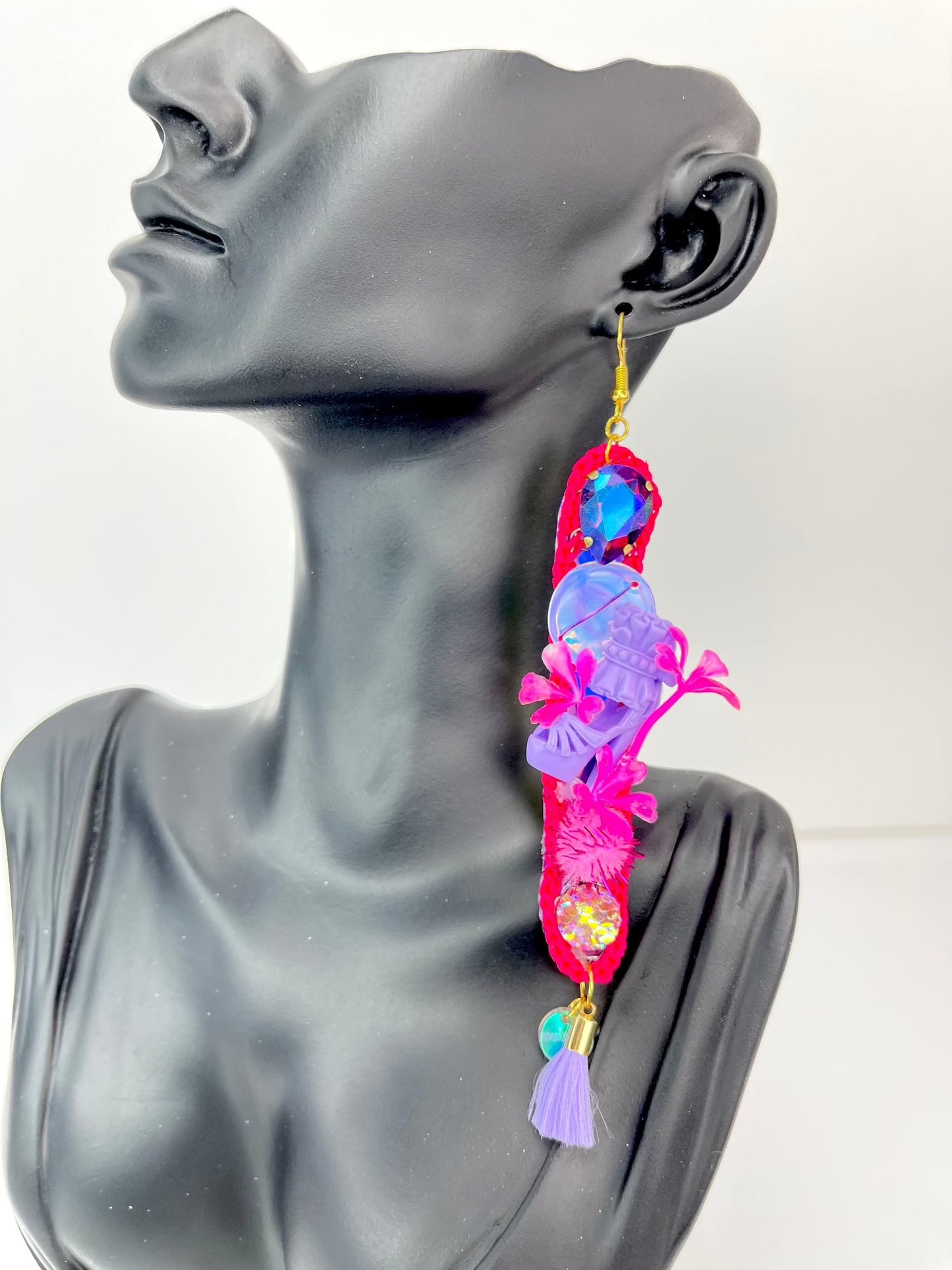 Pink long thin crochet shape, dangle earrings. With purple twinkly stones, iridescent sequins, miniature dolls shoes, flowers, pom poms and lilac tassels. Displayed on a black bust, that is quater human sizes head.