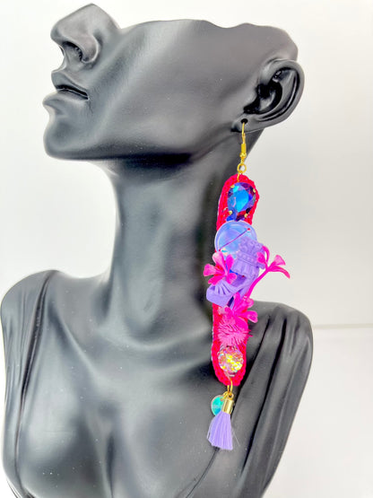 Pink long thin crochet shape, dangle earrings. With purple twinkly stones, iridescent sequins, miniature dolls shoes, flowers, pom poms and lilac tassels. Displayed on a black bust, that is quater human sizes head.