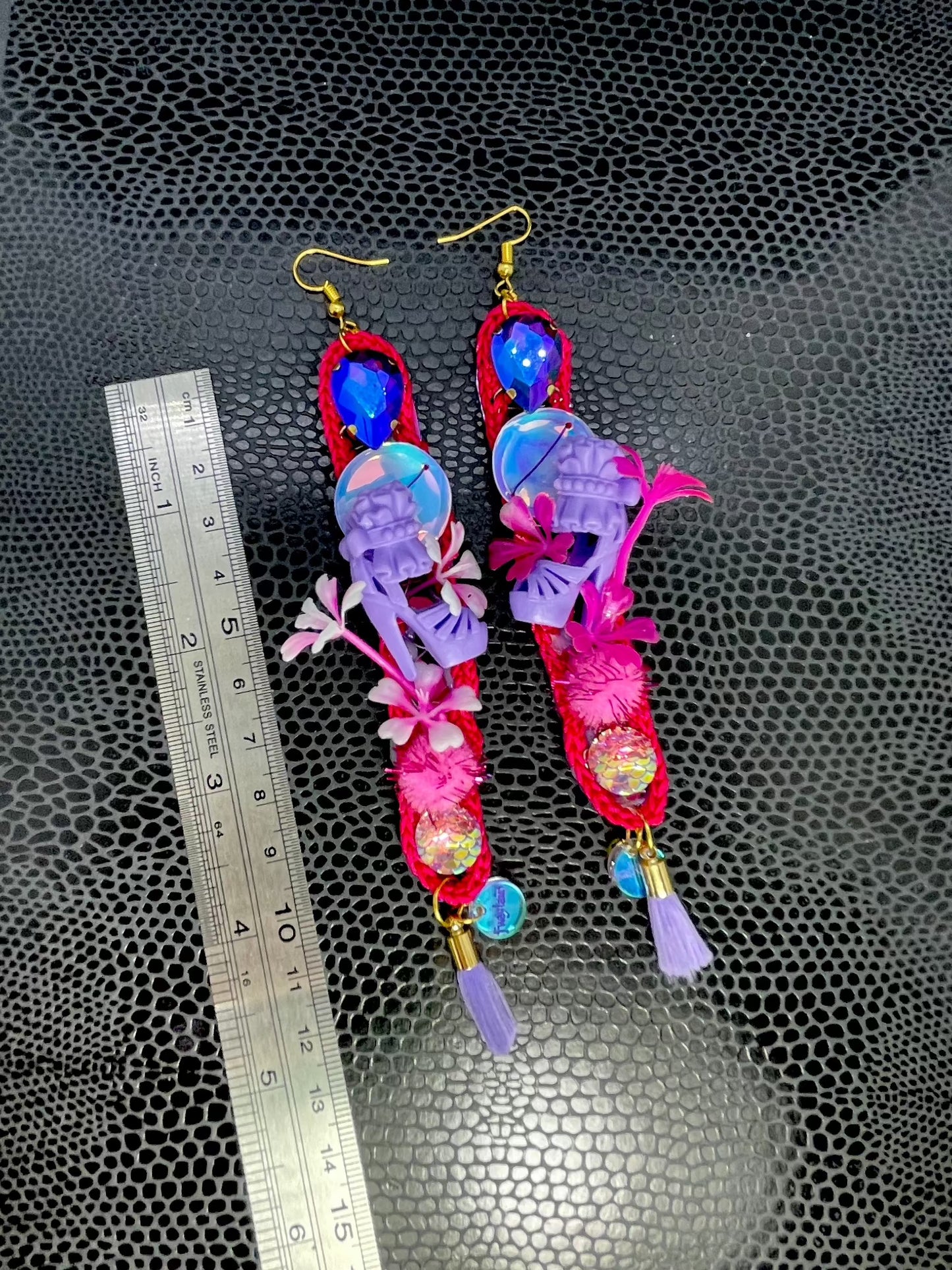 Pink long thin crochet shape, dangle earrings. With purple twinkly stones, iridescent sequins, miniature dolls shoes, flowers, pom poms and lilac tassels. Showing the earrings beside a ruler for scale purposes- they are 12cm long.