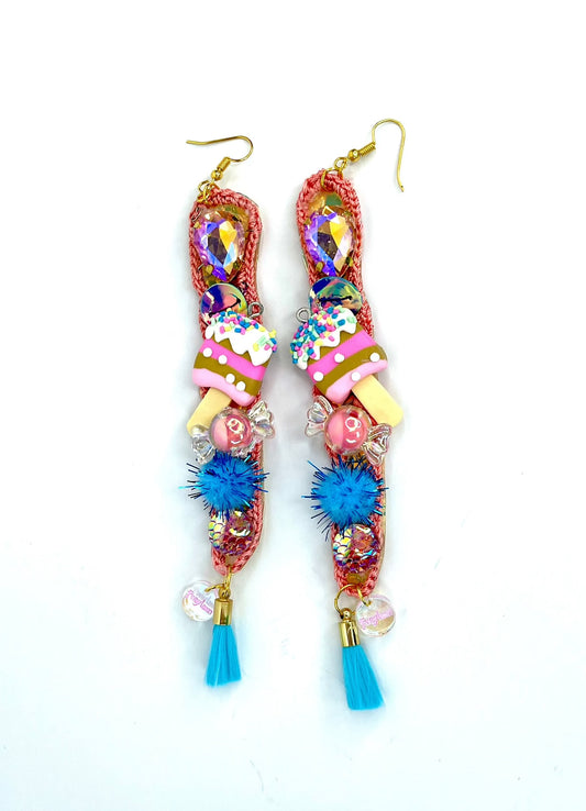 Gold coloured earring parts, with a long skinny crochet oval shape in a salmon pink colour. A large twinkly stone features at the top followed by iridescent sequins, miniature ice cream with sprinkles, sweet beads, baby blue pom poms and tassels